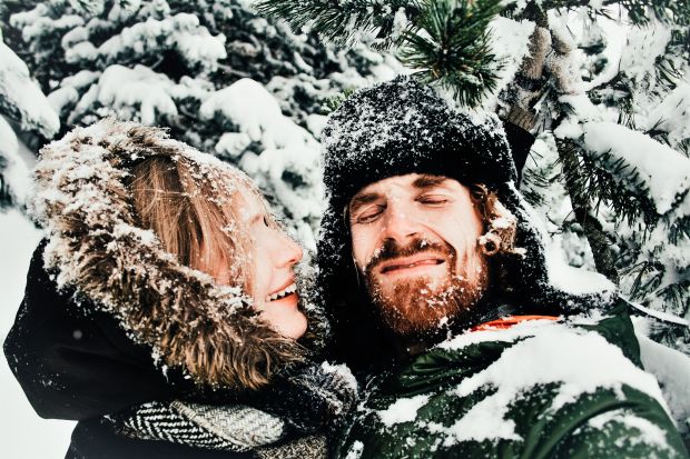 Mann hält eine Frau in einer Schneelandschaft im Arm. Beide sind voll Schnee und lachen.