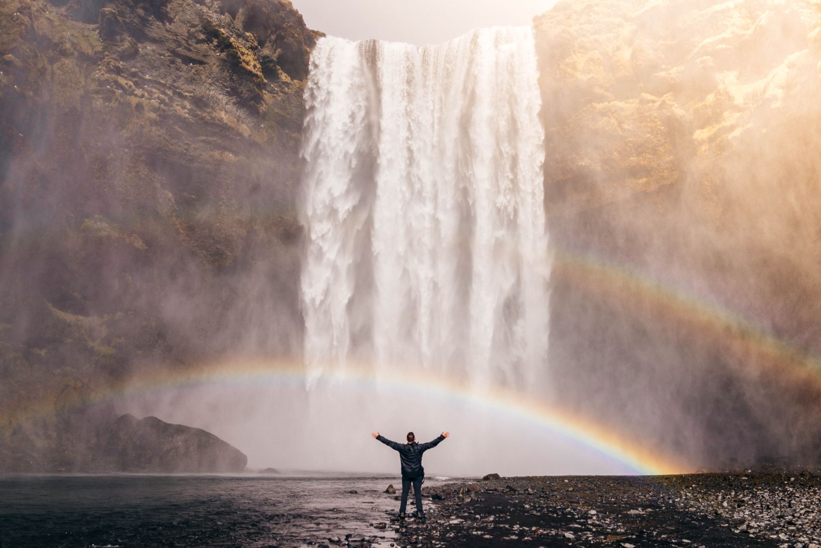 Mann steht mit offenen Armen vor einem Wasserfall in Island. Zwischen ihm und dem Wasserfall ist ein Regenbogen zu sehen.
