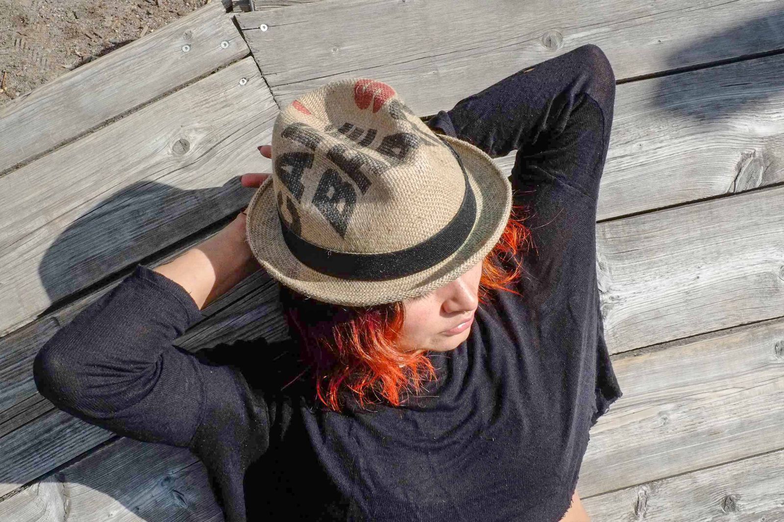 Rotharige, junge Frau liegt auf einem Holzsteg und trägt einen Hut aus einem Kaffeesack